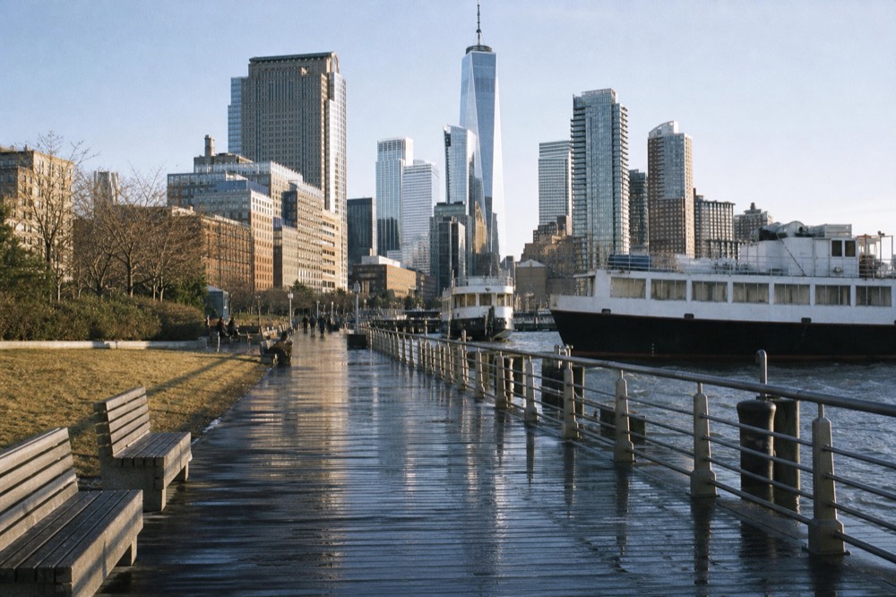 A wide public pier deck with lawn, benches and railings beside moored boats on the Hudson, midday sun and city buildings behind
