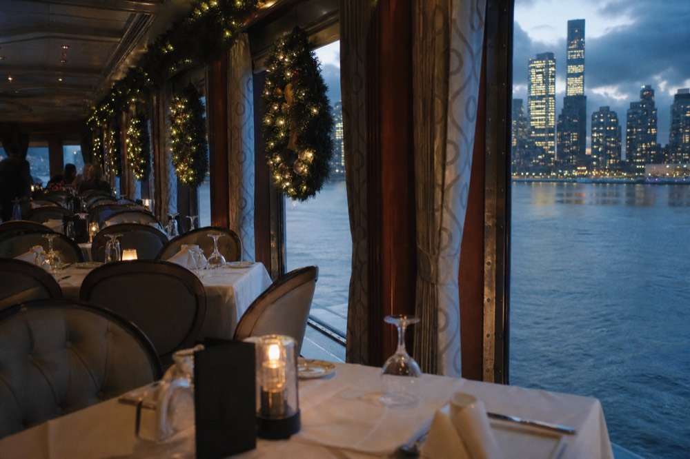 Passengers holding mugs of cocoa inside a warmly lit glass observatory on a small yacht, harbor lights and a bridge visible through the windows at night