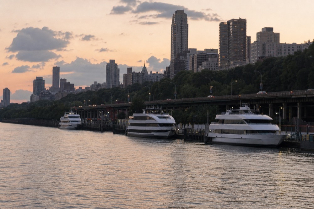 Wide view of a river pier complex at dusk with several excursion vessels moored along a bulkhead and a highway running behind it