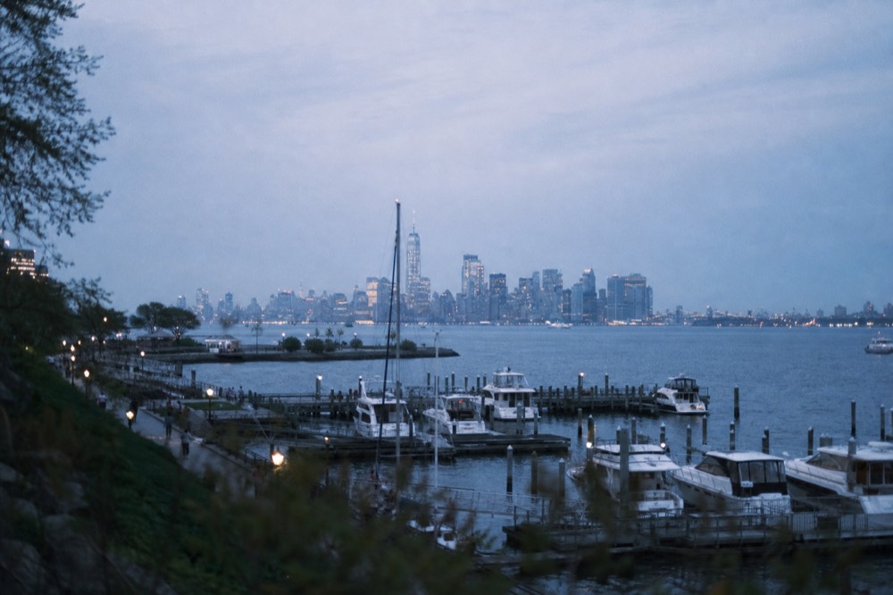 Marina docks in a green waterfront park with the distant Lower Manhattan skyline across open water in hazy afternoon light