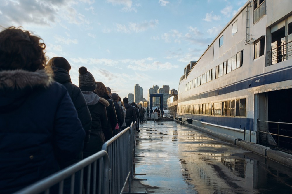 Passengers queuing along a pier walkway beside a moored cruise vessel in late afternoon light, gangway rail in the foreground