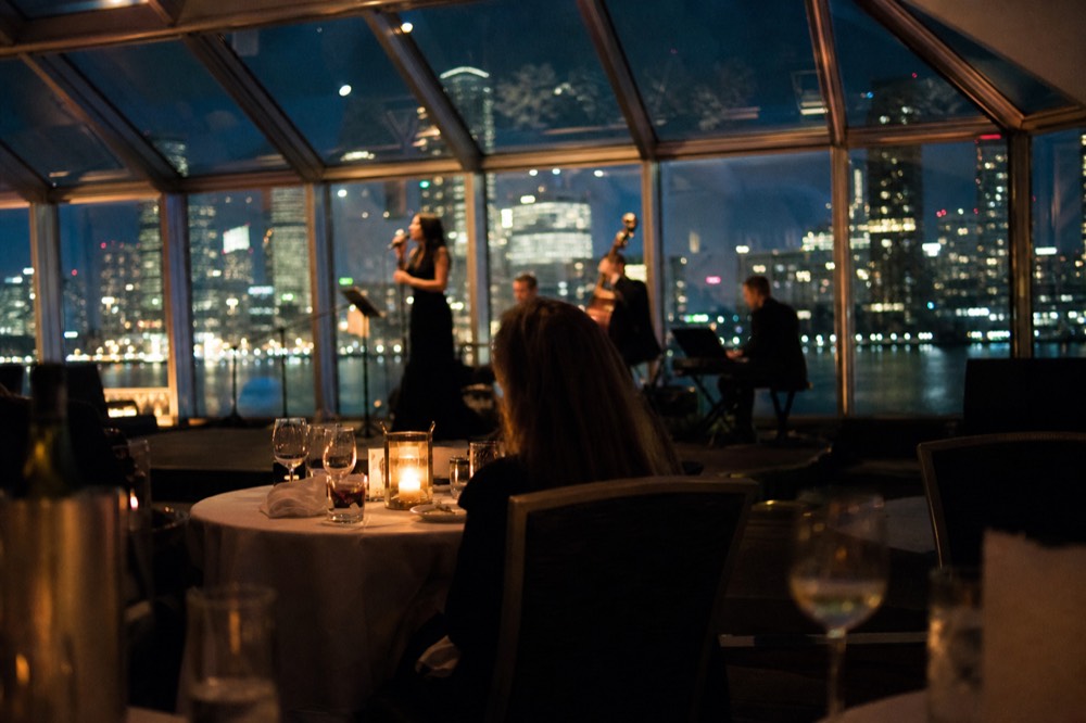 Interior of an all-glass dining room on a cruise vessel with linen-set tables, a singer and small band on a low stage, night skyline through the glass