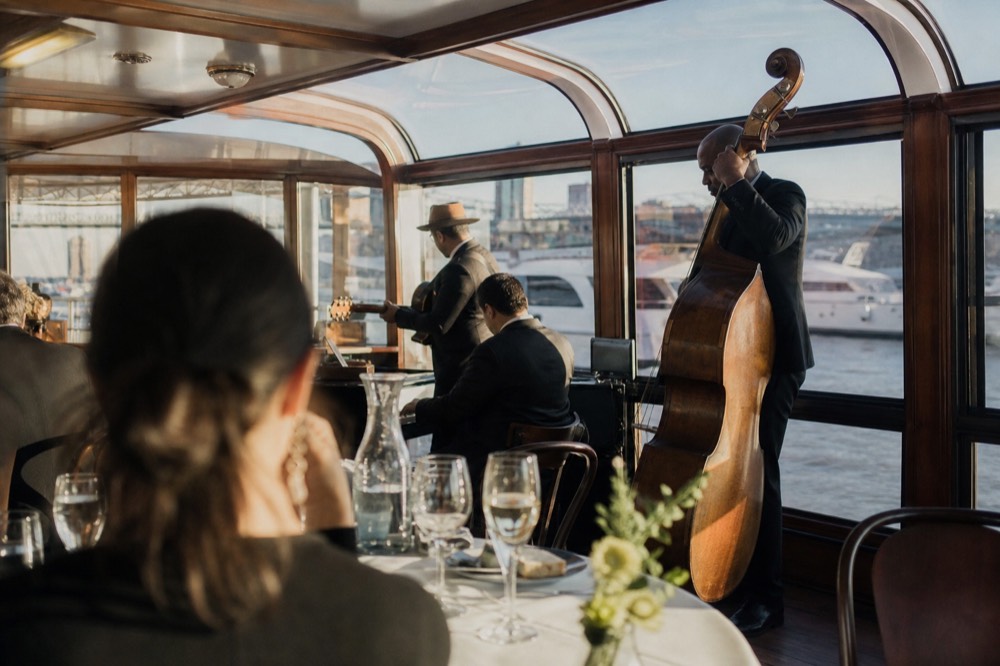 A jazz trio with upright bass playing inside the glass observatory of a small vintage yacht, seated passengers listening with drinks