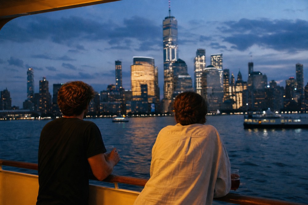 Two people in casual clothes leaning on a boat railing with drinks, city towers rising across the water in soft evening haze