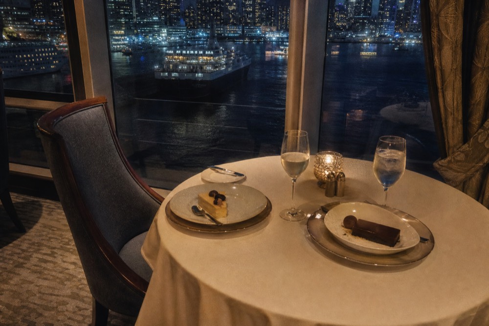A candlelit two-person table by a large cruise ship window with dessert plates and two glasses, city lights reflected in the glass