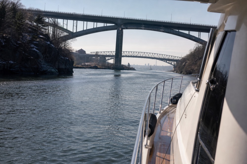 Narrow river passage between rocky banks and bridge spans with a small yacht heading through in flat morning light