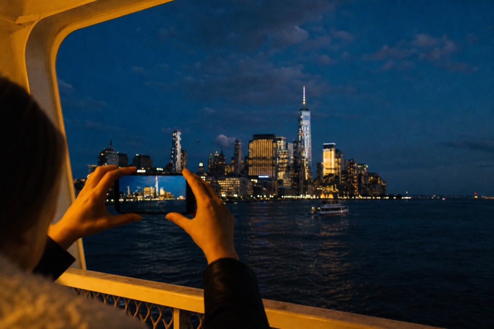 Hands holding a phone up at a boat rail framing the illuminated skyline with the deck rail visible in the lower foreground
