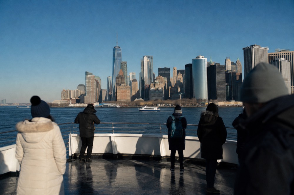 Open upper deck of a cruise vessel in bright daylight with passengers at the rail and the lower Manhattan skyline sharp behind them