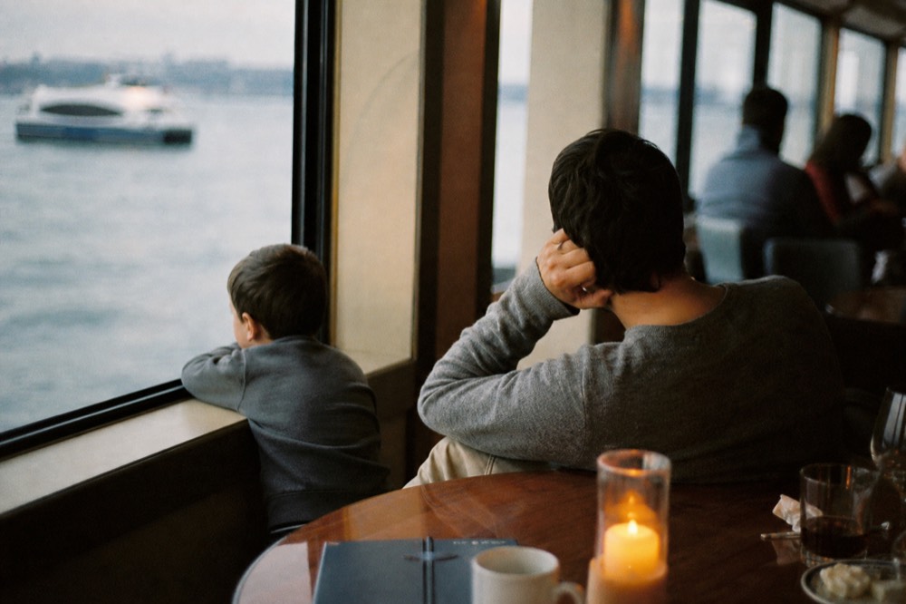 Family group at a round table on a cruise vessel deck, a child looking out of a large window at passing water, casual clothing