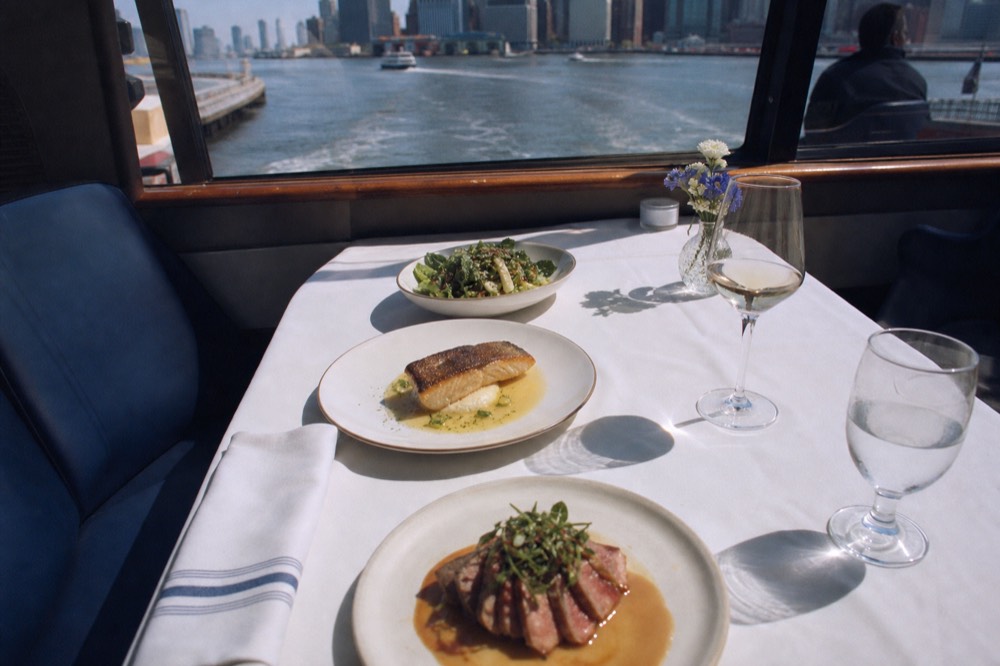 Overhead view of a plated three-course dinner setting on a boat table with a folded napkin, wine glass and city lights blurred beyond the window