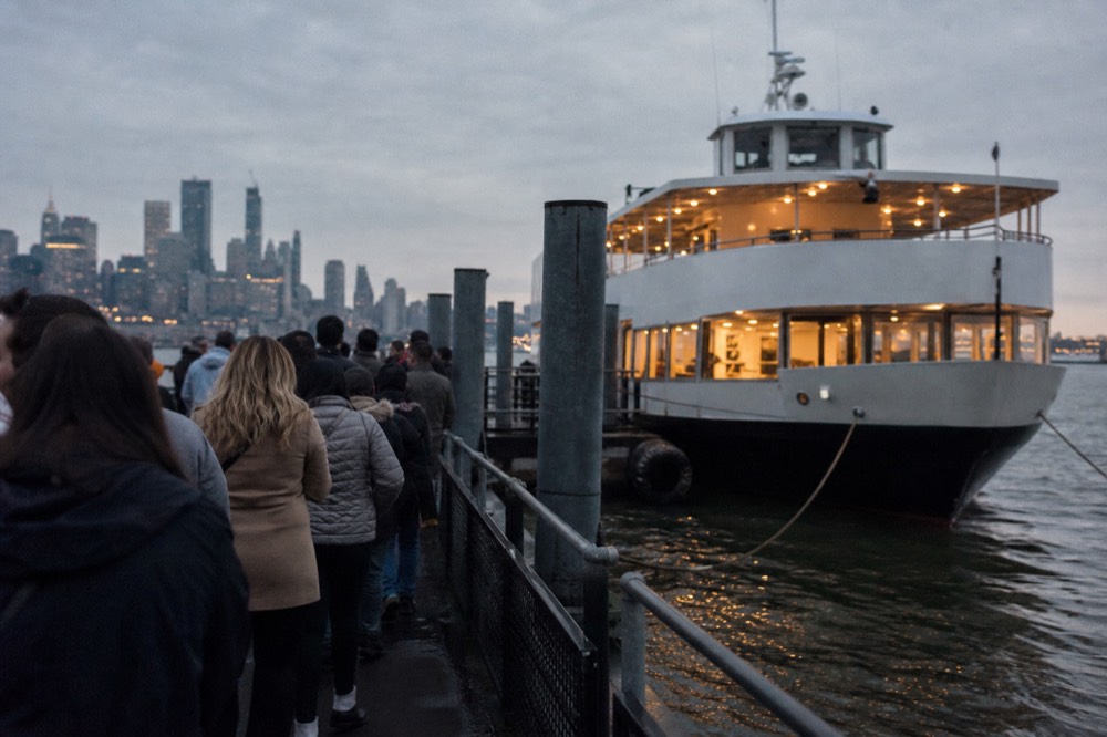 Guests in coats queueing on a Hudson River pier at dusk beside a moored two-deck excursion boat lit from within