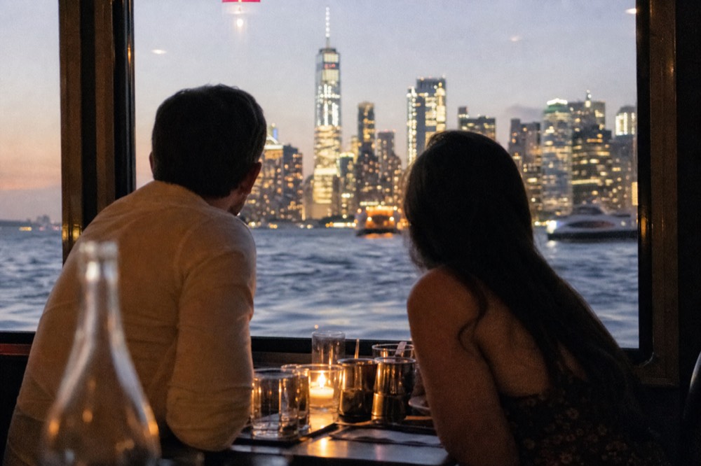 Couple seated at a window table on an evening boat with the Manhattan skyline glowing behind them, faces turned away toward the view