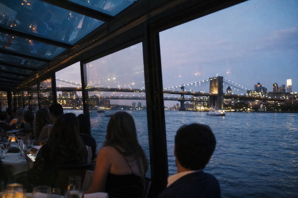 Row of guests in cocktail dresses and jackets seated at linen-covered tables inside a fully glazed boat saloon at dusk