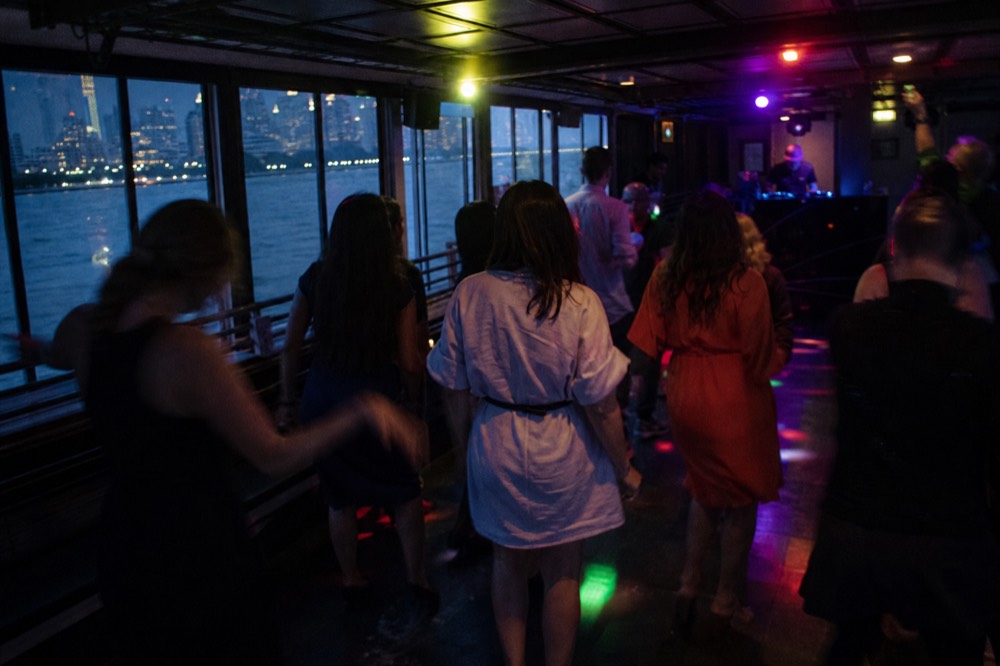 Guests dancing on a boat's interior floor under coloured lights with a DJ booth to one side and windows showing city lights