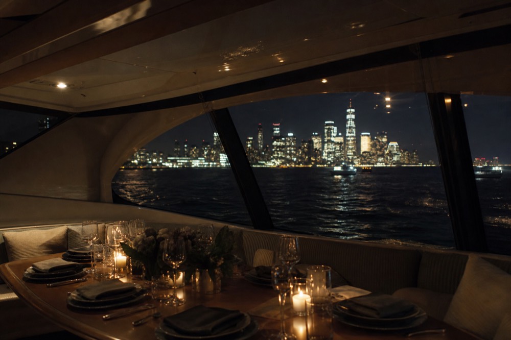Small yacht saloon with a set table for six, low warm lighting and dark water visible through the side windows